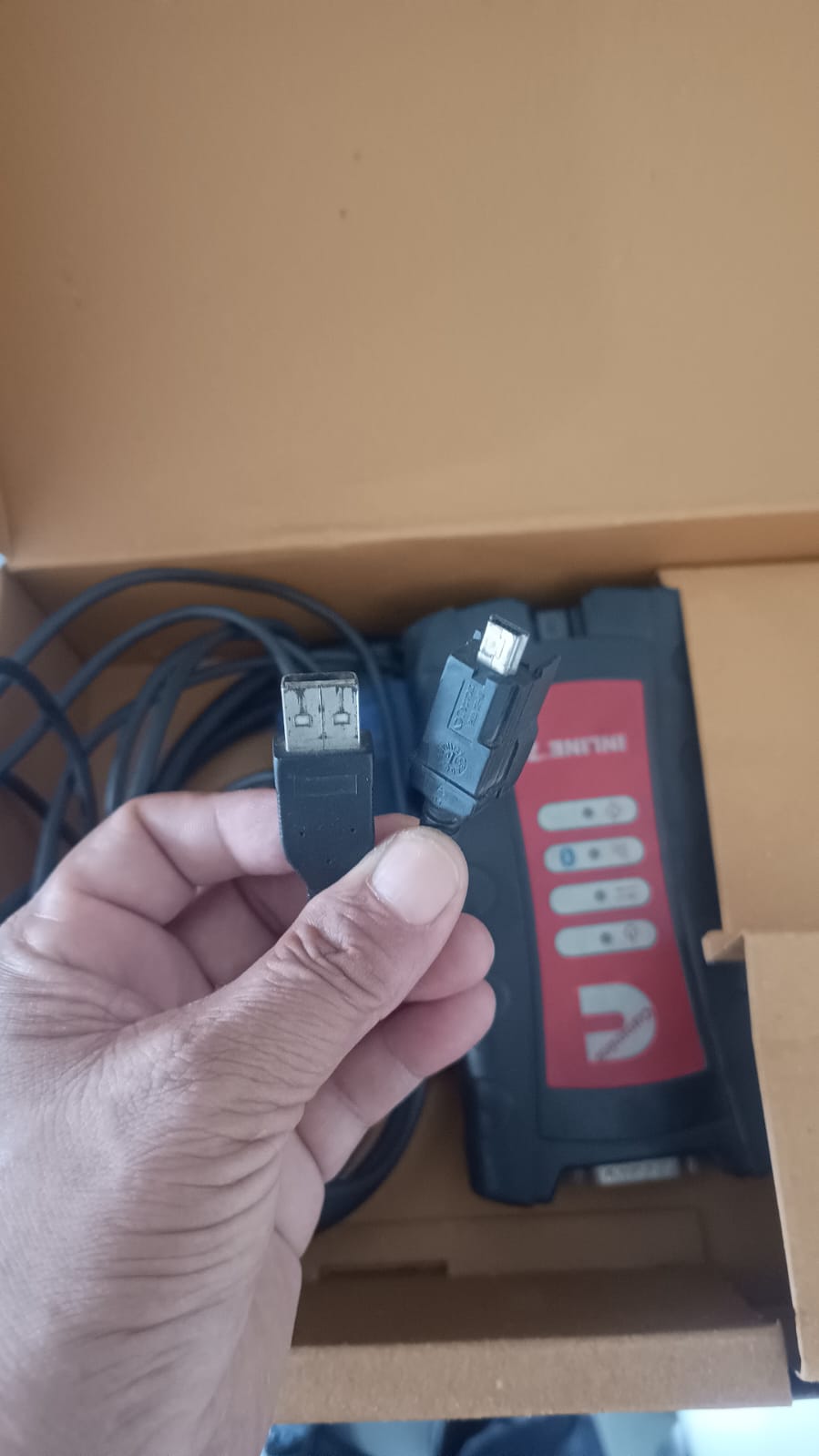 Person holding a red and black power tool battery with a charger cable in front of an open cardboard box.